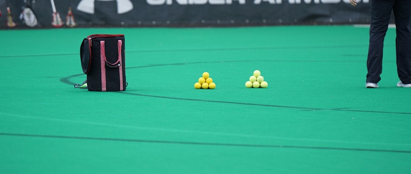 A sports field features a black and pink sports bag, two pyramids of yellow balls, and a portion of a person in dark athletic gear. The background displays a banner with the Under Armour logo.