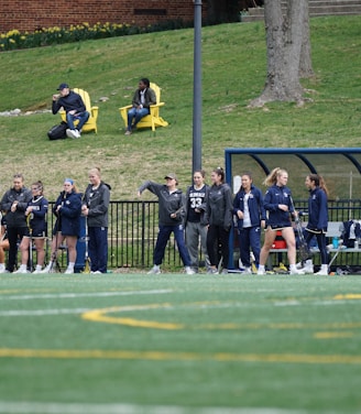 A group of female athletes, some in sports uniforms and others in jackets, are gathered along the sideline of a sports field. They seem to be in a team environment, possibly preparing for or observing a game. In the background, there are a few people sitting on yellow chairs on a grassy incline, and a building with a brick facade.