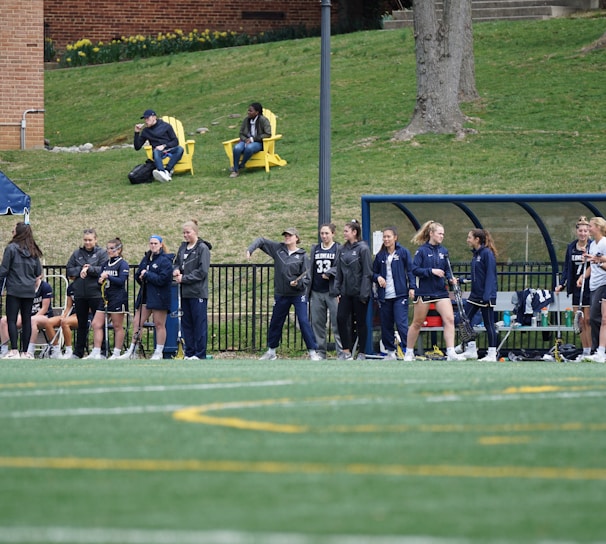 A group of female athletes, some in sports uniforms and others in jackets, are gathered along the sideline of a sports field. They seem to be in a team environment, possibly preparing for or observing a game. In the background, there are a few people sitting on yellow chairs on a grassy incline, and a building with a brick facade.