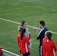 A group of people wearing sports attire are gathered on a grassy field. Some individuals are dressed in red jackets, while others are in turquoise and navy blue. A coach appears to be giving instructions. Sports equipment, such as water bottles and a bench with towels, can be seen nearby.