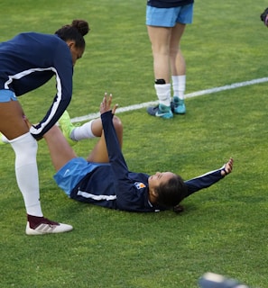 A group of athletes in sportswear are on a green soccer field. Two individuals are interacting closely; one helps the other who is lying on the grass. Nearby, other athletes are stretching.