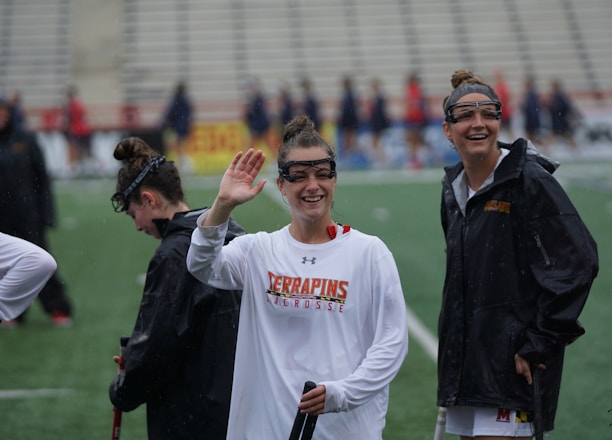 Several athletes in sports attire are on an outdoor sports field, interacting and smiling. One individual in the foreground waves with a friendly expression, while another stands beside her, also smiling. They wear protective goggles and gear associated with lacrosse. The background features indistinct figures in dark clothing against a backdrop of stadium seating.