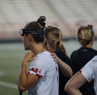 Several people wearing sports attire and protective eyewear are gathered on a sports field. They appear to be engaged in a discussion or briefing, possibly during a break from a game or practice. The background shows a blurred stadium seating, indicating an outdoor setting.