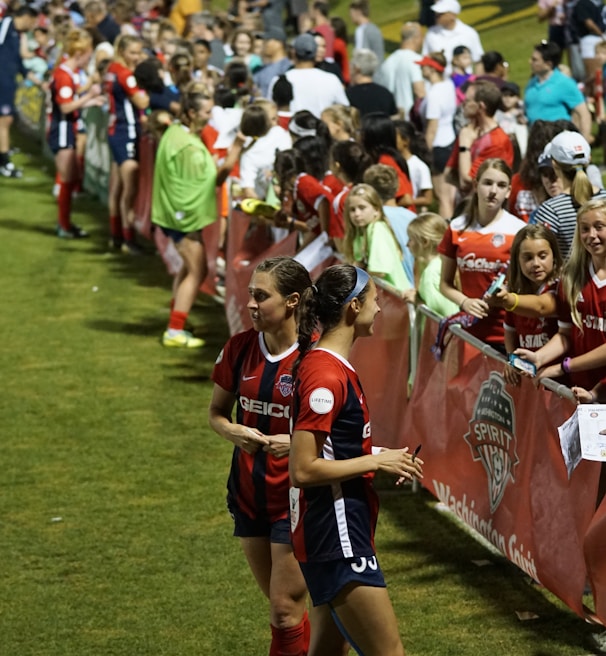 The image features a lively gathering of young soccer players and fans next to a field. Several female athletes in red and blue uniforms interact with an enthusiastic crowd standing behind a barrier. Some people in the crowd are holding objects, like pens and paper, possibly for autographs.