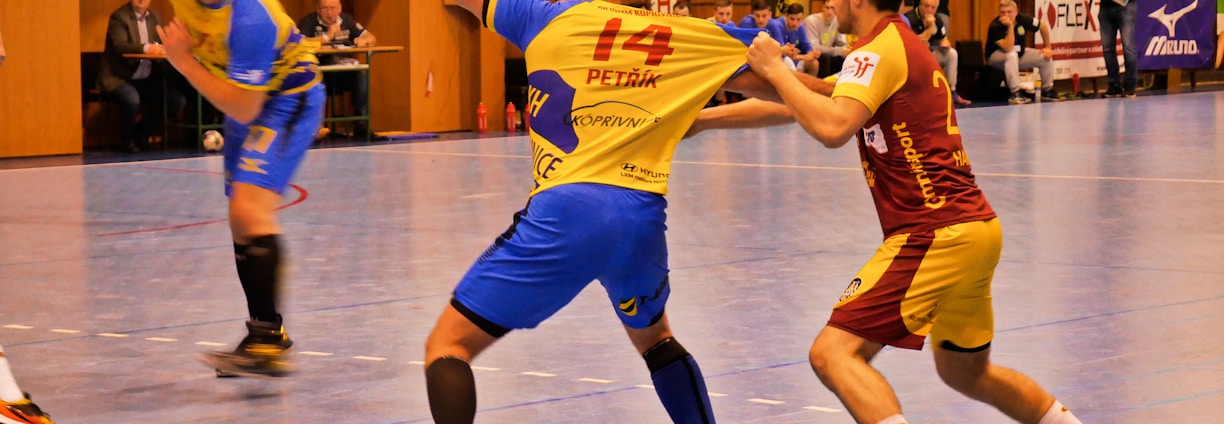 A dynamic indoor sports scene with players in motion on a court. Two athletes in blue and yellow uniforms are engaged in an intense handball game, with one player gripping the other's jersey. The background shows other players, several seated people, and advertisements on the walls.